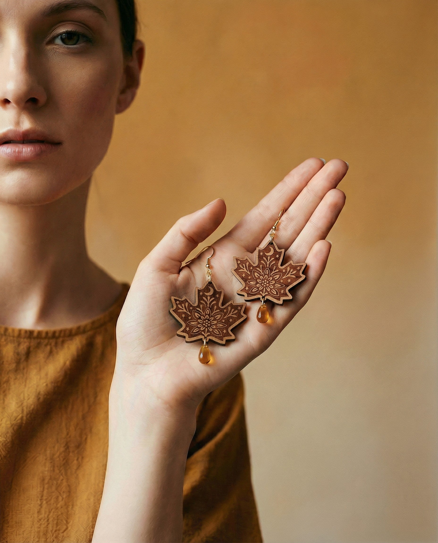 Hand holding pair of engraved maple leaf wood dangle earrings with amber Czech glass teardrop beads and 14k gold hooks.