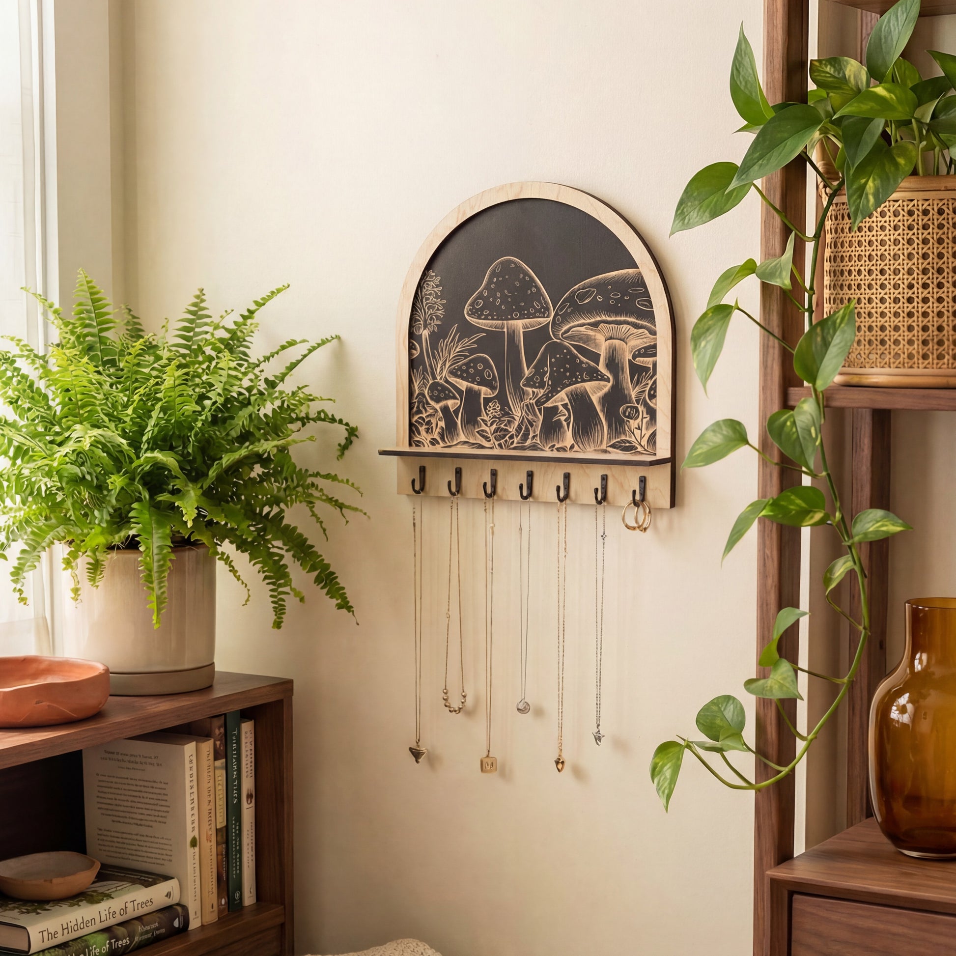 Arched wooden wall organizer with engraved mushroom artwork and black hooks, used as a key holder and jewelry hanger with necklaces, styled with plants and shelves.