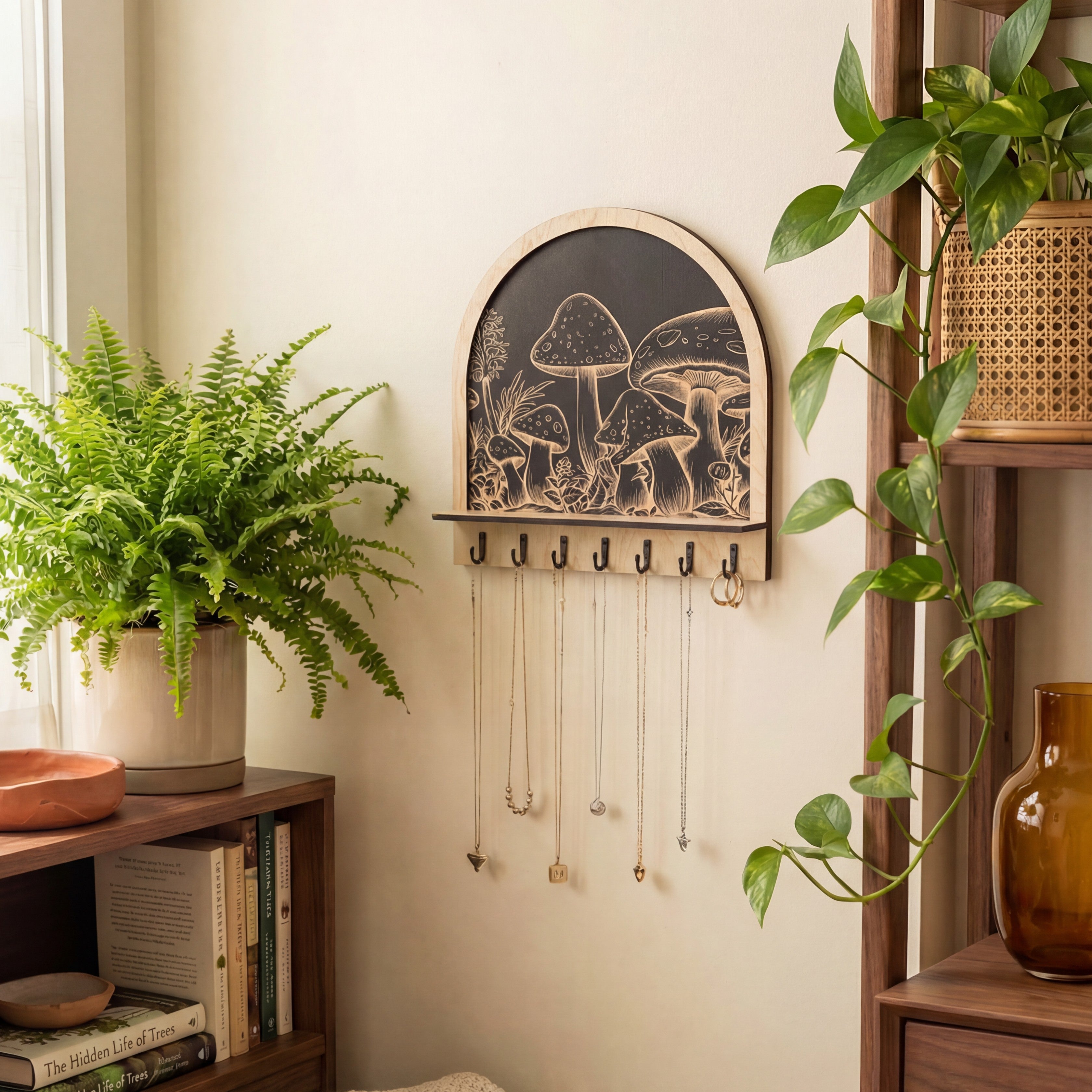 Arched wooden wall organizer with engraved mushroom artwork and black hooks, used as a key holder and jewelry hanger with necklaces, styled with plants and shelves.