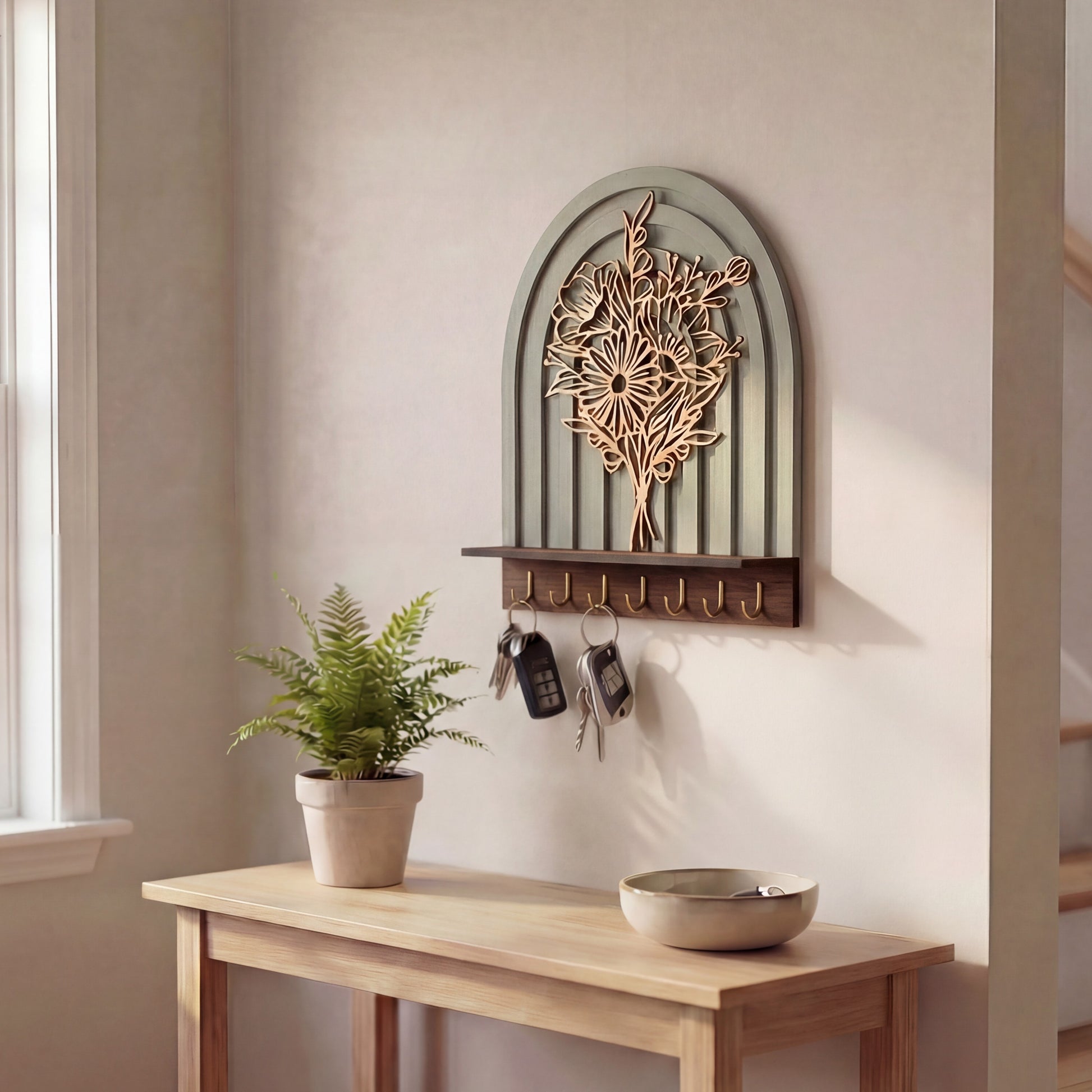 Arched wall-mounted key holder with layered floral botanical design, wood shelf, and brass hooks holding keys in an entryway.