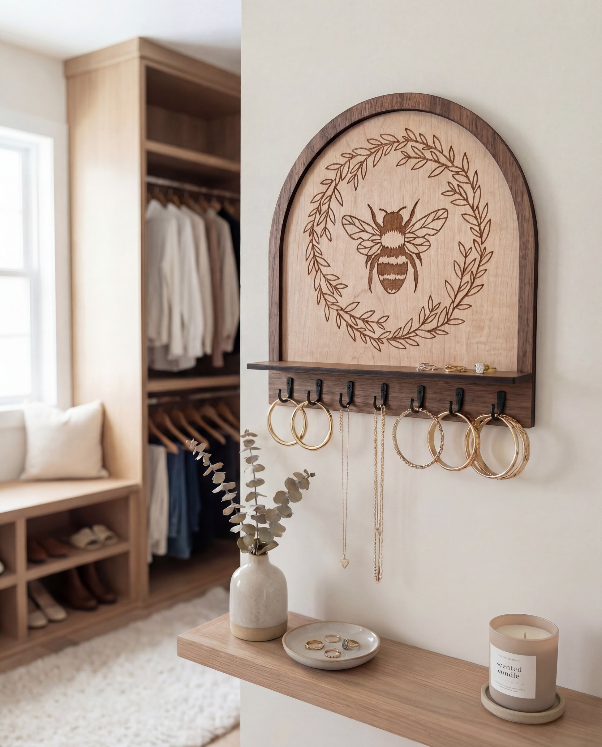Maple wood bee wall organizer key holder with engraved honeybee wreath design and black hooks, shown holding hoop earrings and necklaces on a neutral wall with a styled shelf, vase, and closet entryway background
