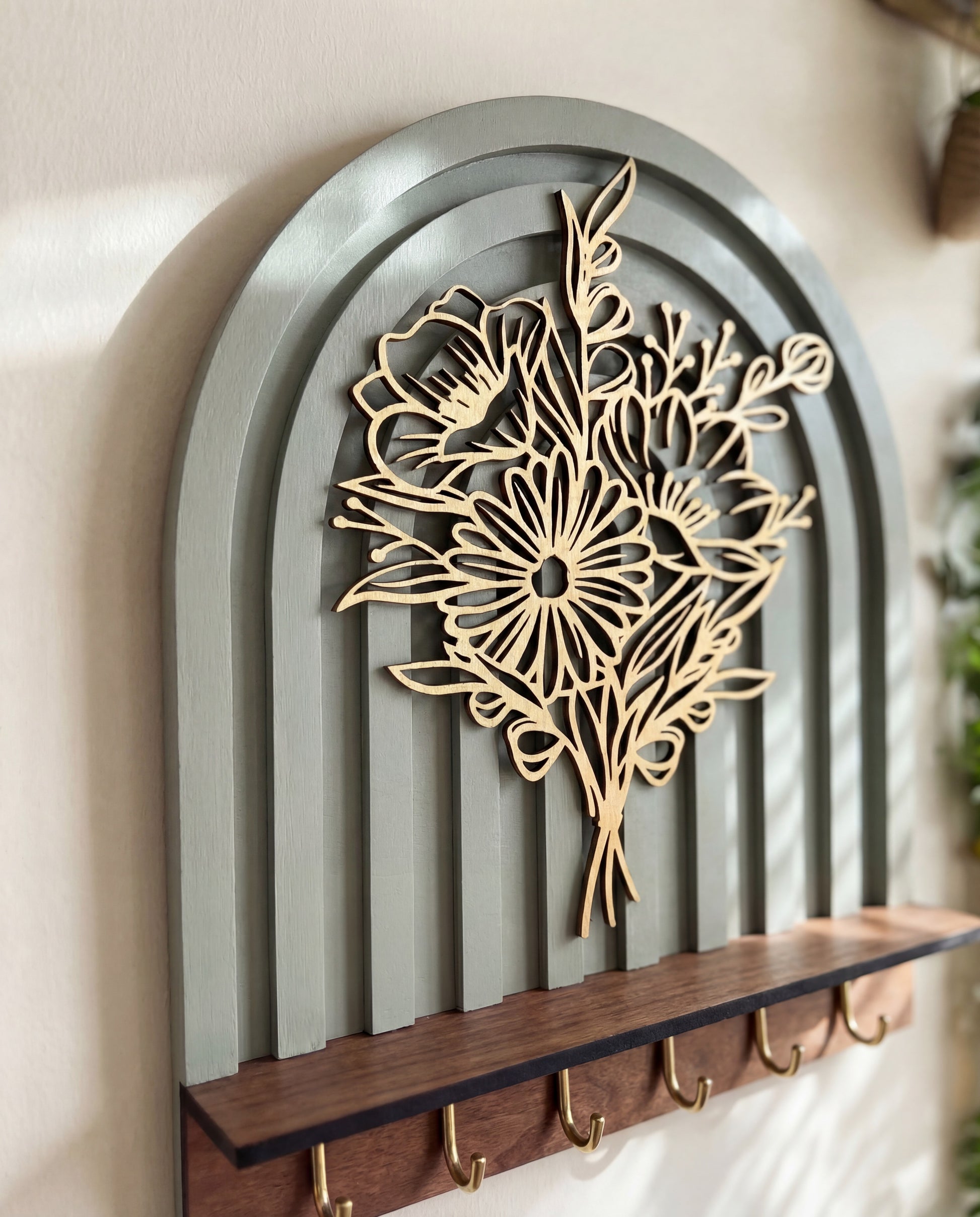 Close-up of an arched wall key holder featuring a layered botanical floral design, dark wood shelf, and brass hooks.