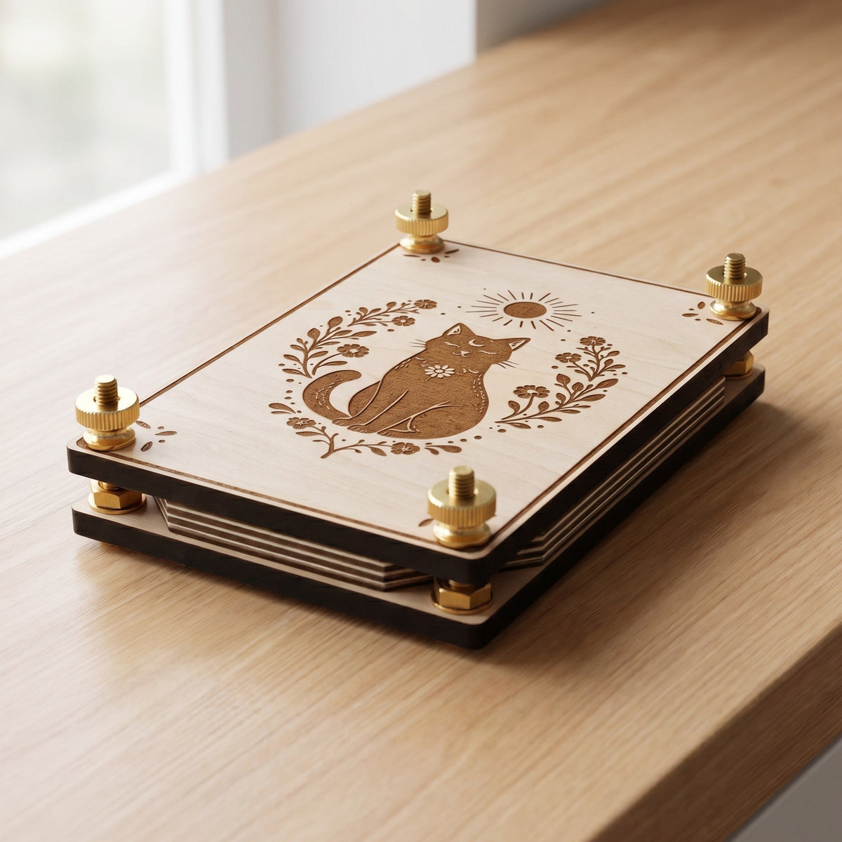 Handcrafted wooden flower press featuring an engraved black cat, botanical florals, and sun motif. Angled side view highlights the brass thumb screws, layered pressing boards, and blotting paper, resting on a light wooden table in natural sunlight.