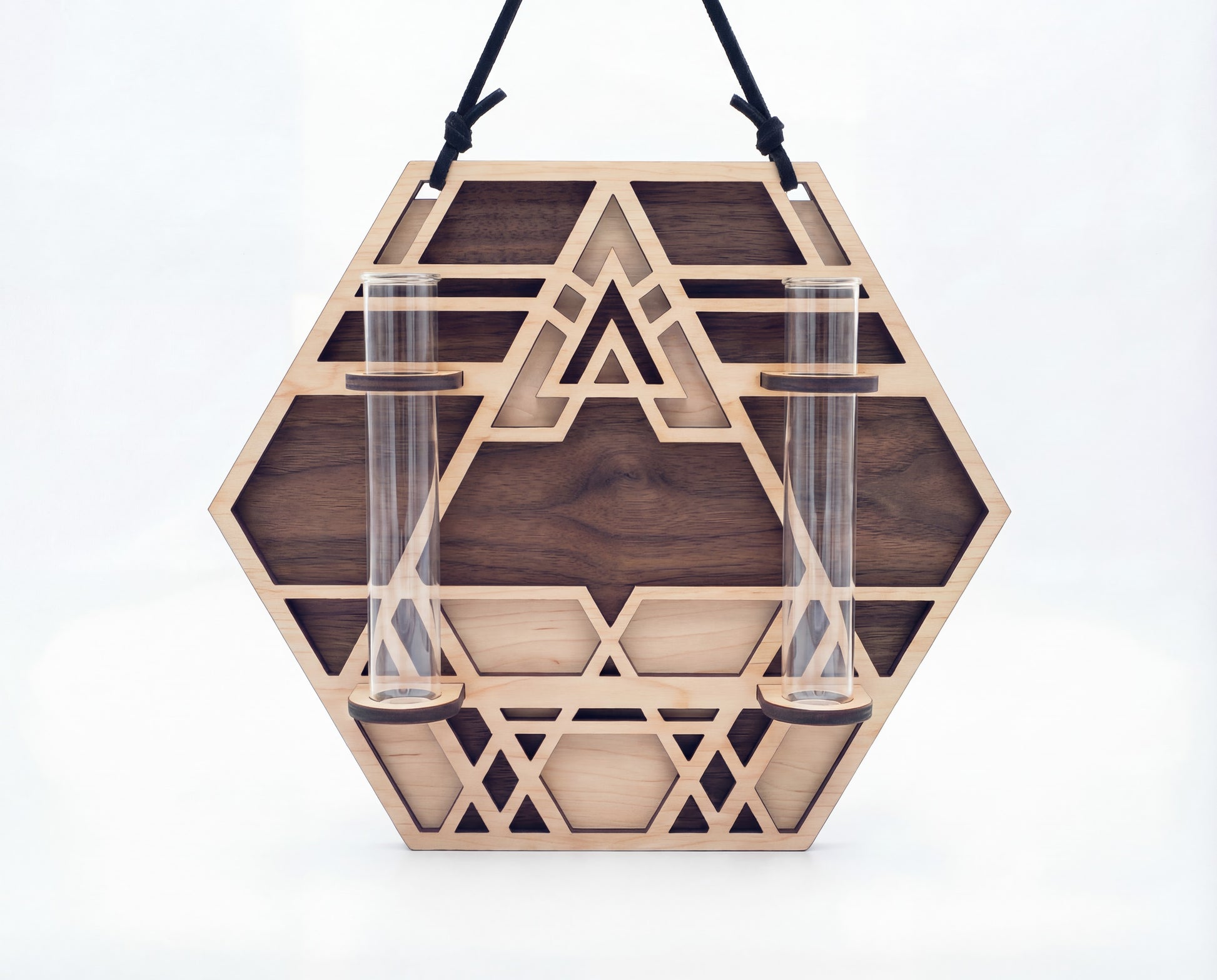 Front view of a geometric hexagon wall propagation station made from layered maple wood with a walnut-toned back layer and sacred geometry cutout design, hanging from a black suede strap and holding two clear 6.25-inch by 1-inch glass vials on a white background.