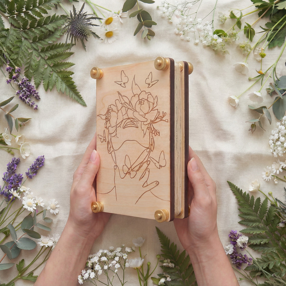 Hands holding a handmade 4x6 maple wood flower press with brass knurled thumb nuts, engraved woman line art with peonies and butterflies, surrounded by fresh wildflowers and greenery on linen.