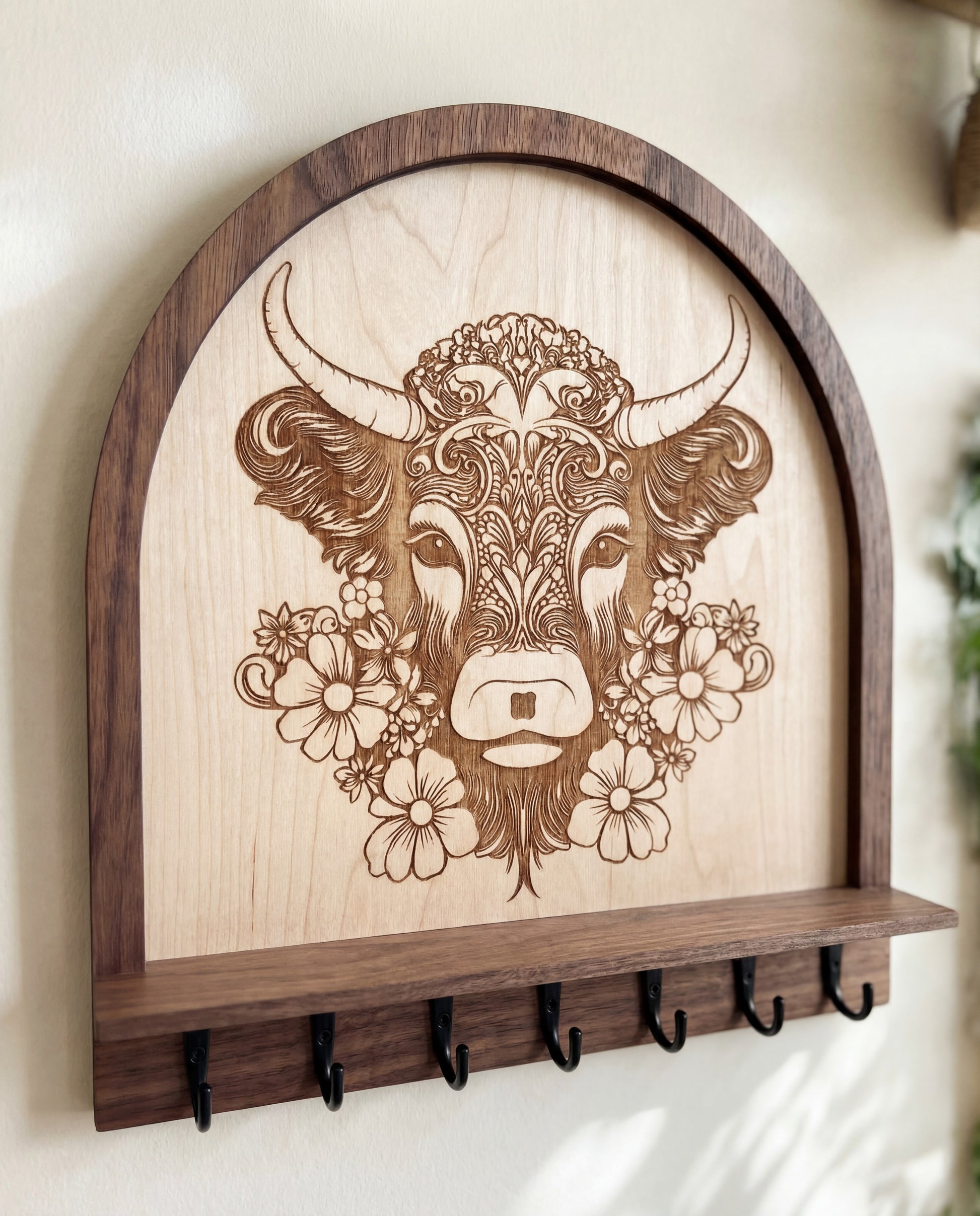 Close-up of a laser engraved floral highland cow design on a wood wall key and jewelry holder with shelf and black metal hooks, rustic farmhouse entryway organizer.
