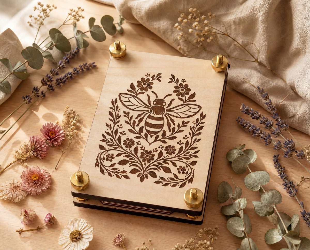 A rectangular flower press with a laser-engraved bee and floral design, shown in a styled flat-lay with dried flowers, eucalyptus, and linen fabric.