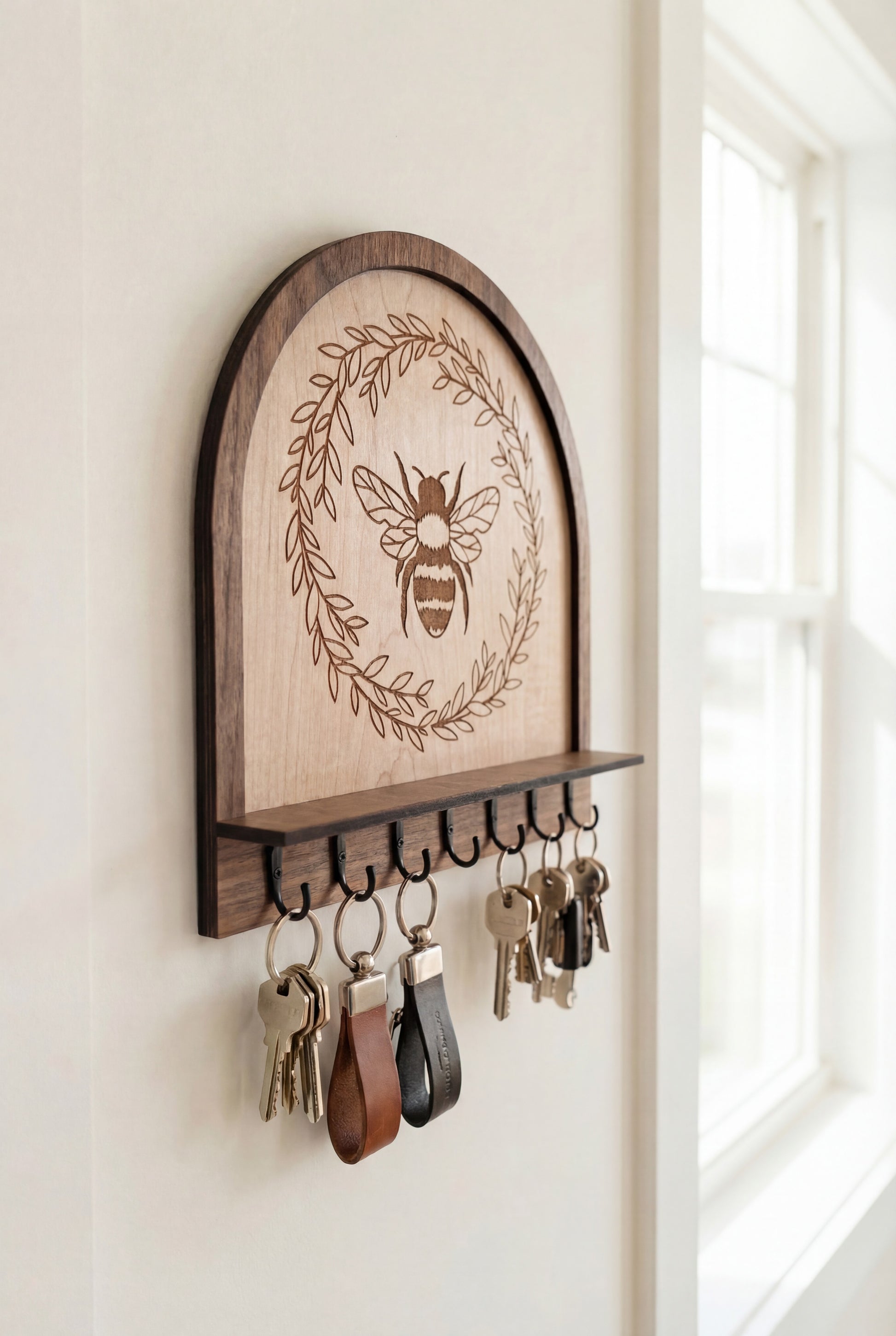 Maple wood bee wall organizer key holder with engraved honeybee wreath design and black metal hooks, shown holding multiple sets of keys on key rings in a bright entryway near a window.