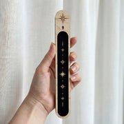 Hand holding a slim wooden incense holder with a black inlay and gold celestial star design.