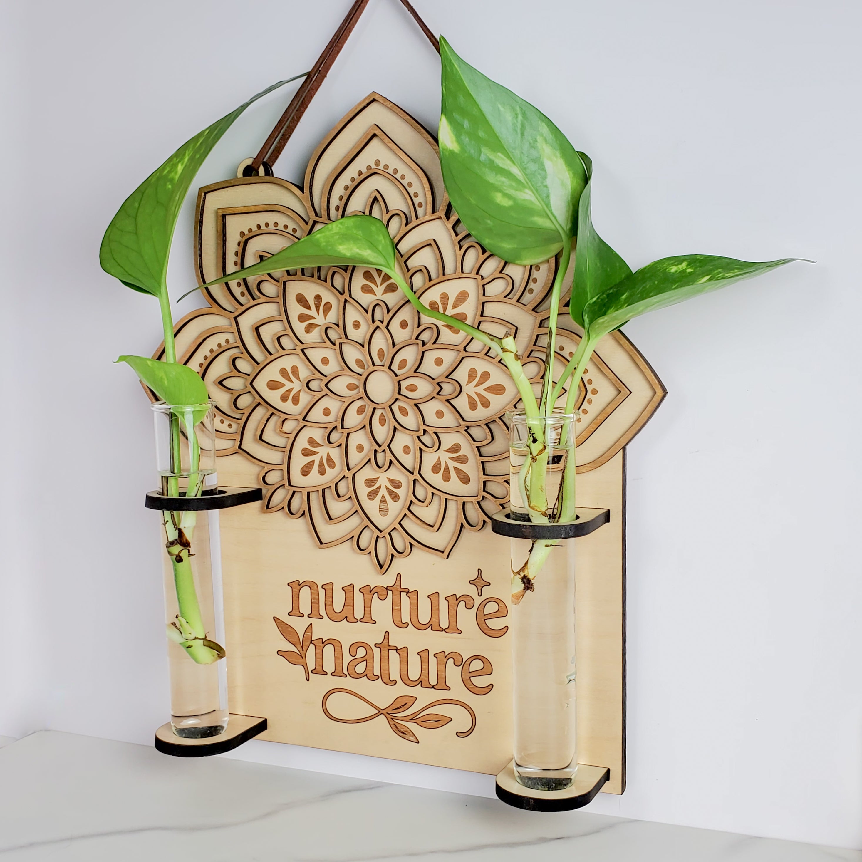 Close-up front view of a layered maple wood mandala wall propagation station with “nurture nature” engraving, suede hanging strap, and two 6.25-inch by 1-inch clear glass vials holding pothos cuttings in water on a white background.
