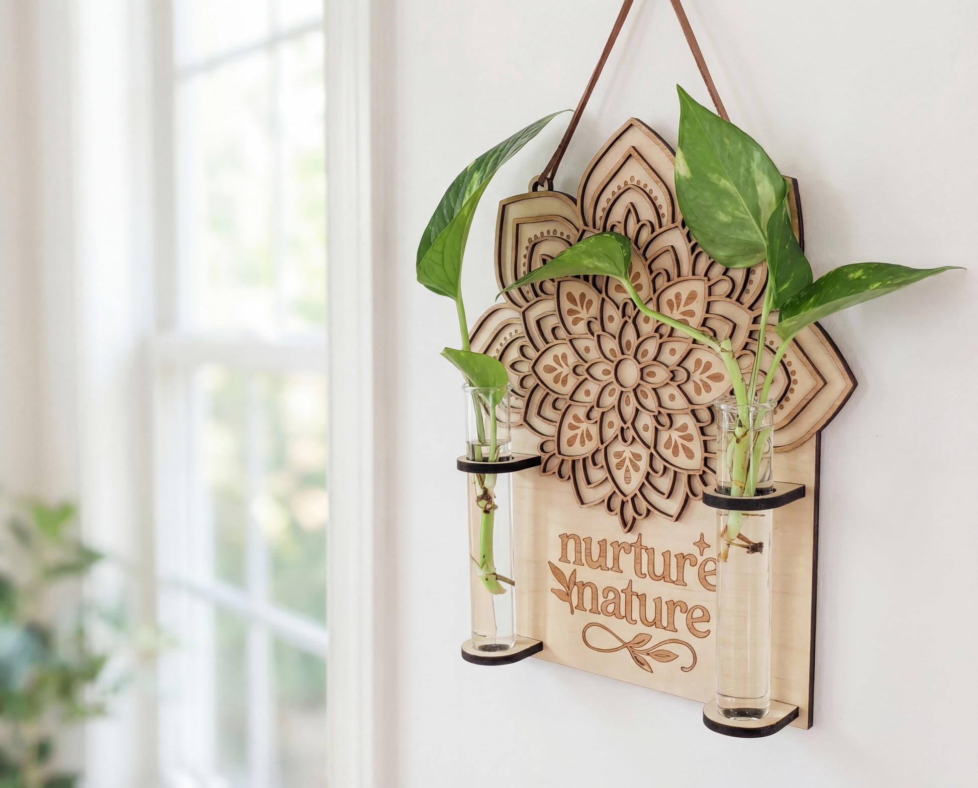 Laser-cut mandala wall propagation station with “nurture nature” engraving, made from layered maple wood and hanging from a suede strap, featuring two clear glass vials holding pothos cuttings in water beside a bright window as boho plant wall decor.