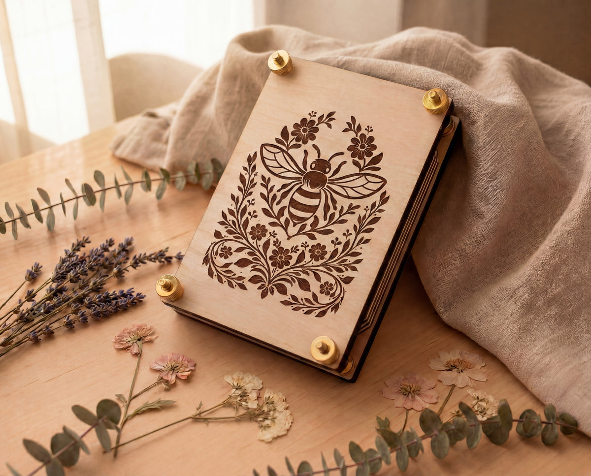 A wooden flower press with a laser-burned bee and floral design on the cover, secured with brass corner brackets, resting on a wooden table with a beige linen cloth and dried flowers around it.