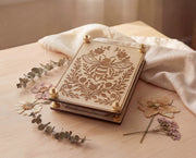 Angled view of a 4x6 engraved wooden flower press kit with a bee and botanical floral design, made from 5.2mm premium maple MDF core board and assembled with brass hardware and knurled thumb nuts, styled on a light wood surface with dried pressed flowers, eucalyptus sprigs, and a soft linen cloth in warm natural light.