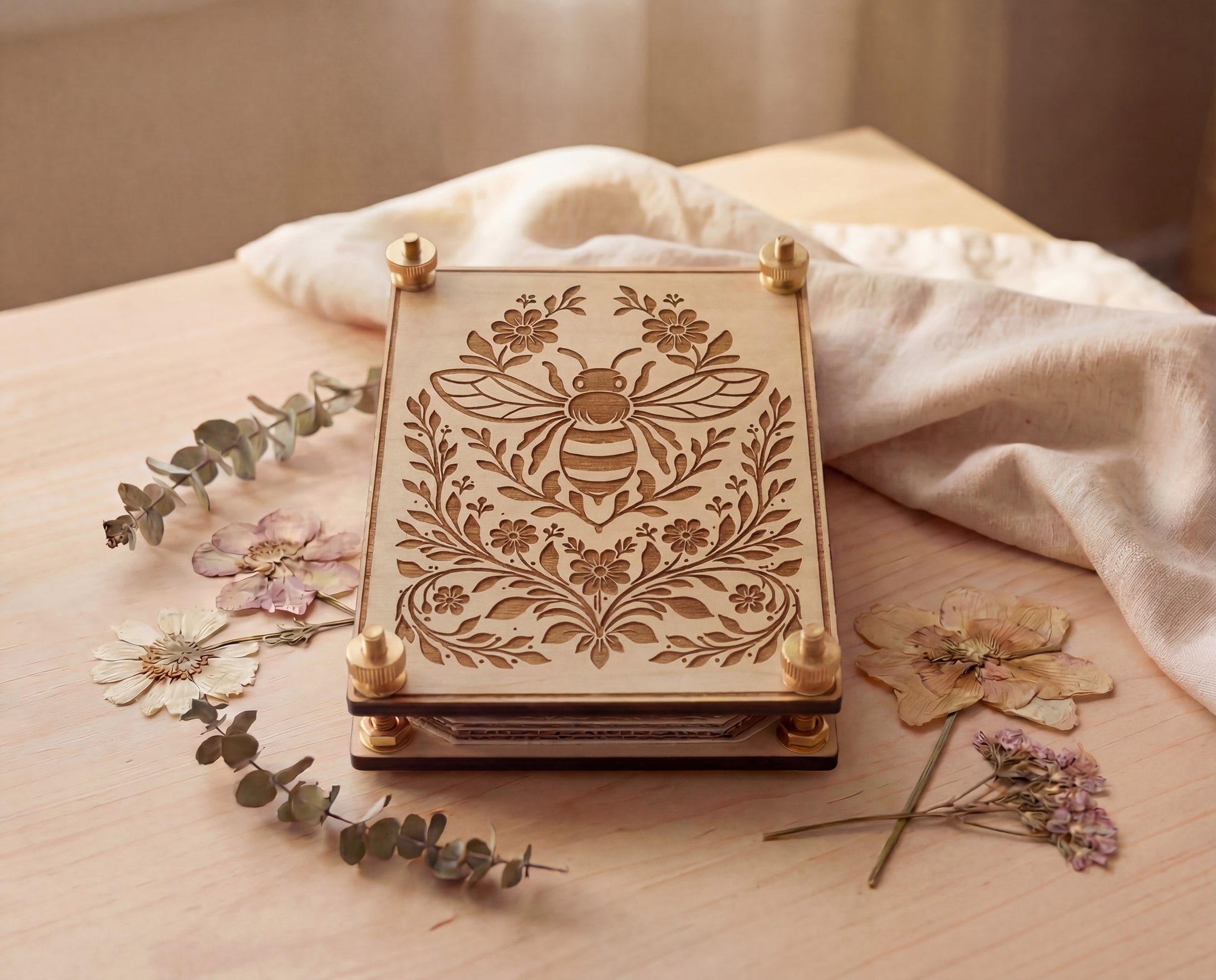 Front view of a 4x6 engraved wooden flower press kit featuring a bee and botanical floral design, made from 5.2mm premium maple MDF core board with brass hardware and knurled thumb nuts, shown stacked with pressing layers on a light wood tabletop beside dried pressed flowers, eucalyptus sprigs, and a soft linen cloth in warm natural light.