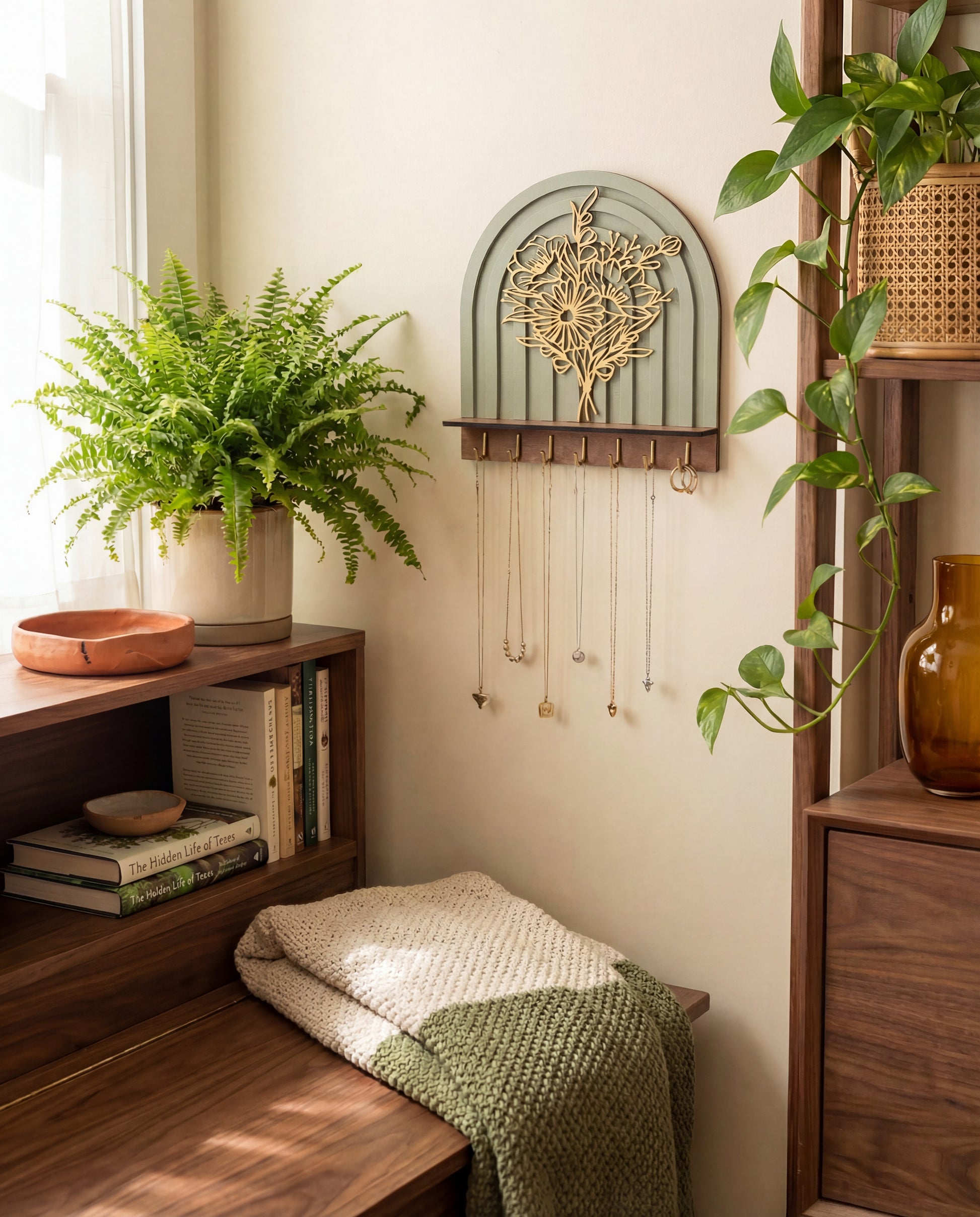 Arched wall-mounted jewelry organizer with layered floral botanical design, wood shelf, and brass hooks holding necklaces in a cozy boho room.