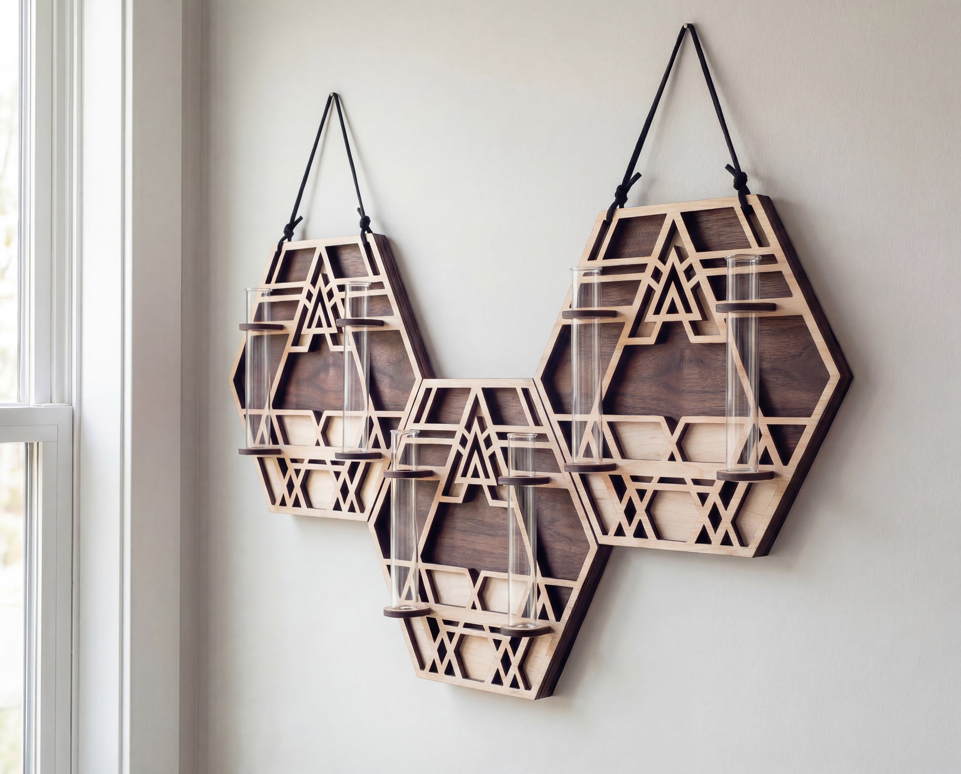 
XL trio geometric wall propagation station made from layered maple wood with walnut-toned back layers and sacred geometry cutout design, hanging from black suede straps and holding six clear glass vials on a white wall near a window.