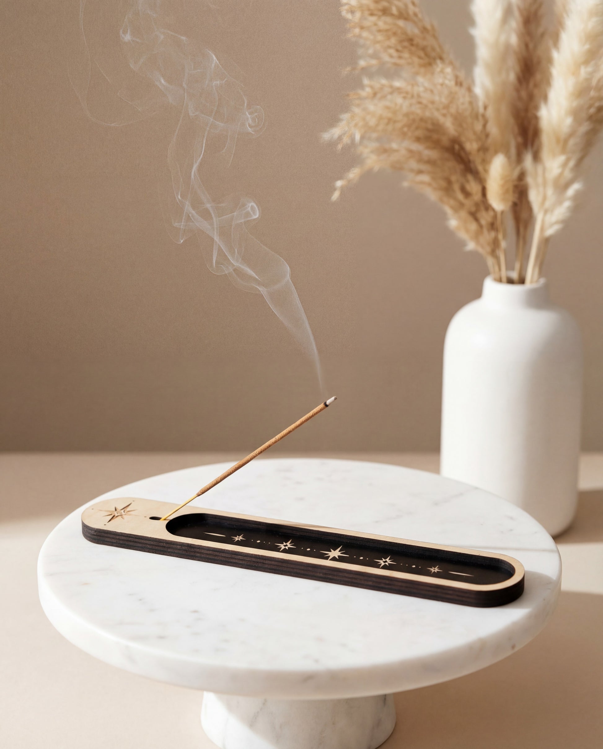 Lit incense stick in a wooden celestial incense holder with black inlay and star design on a marble tray, with smoke rising beside a white vase.