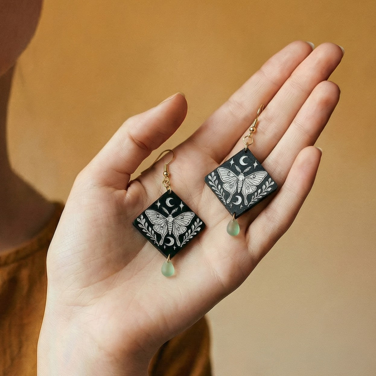 Hand holding pair of engraved moth diamond wood dangle earrings with crescent moon design, mint Czech glass teardrop beads, and 14k gold hooks.