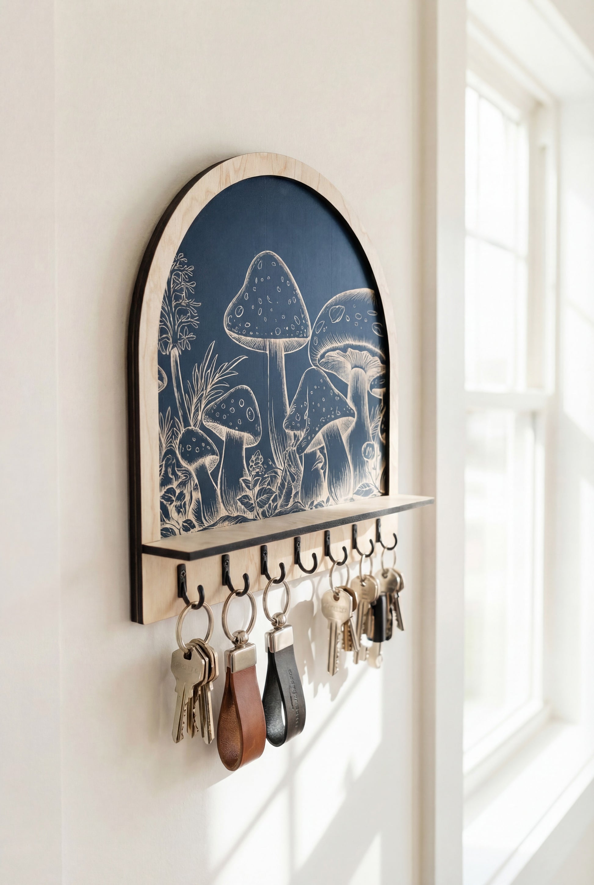 Close-up of an arched wooden wall key holder with engraved mushroom artwork and black hooks holding keyrings, photographed in bright natural window light.