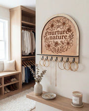 Arched wooden wall jewelry organizer with “nurture nature” floral engraving, holding hoop earrings and necklaces.