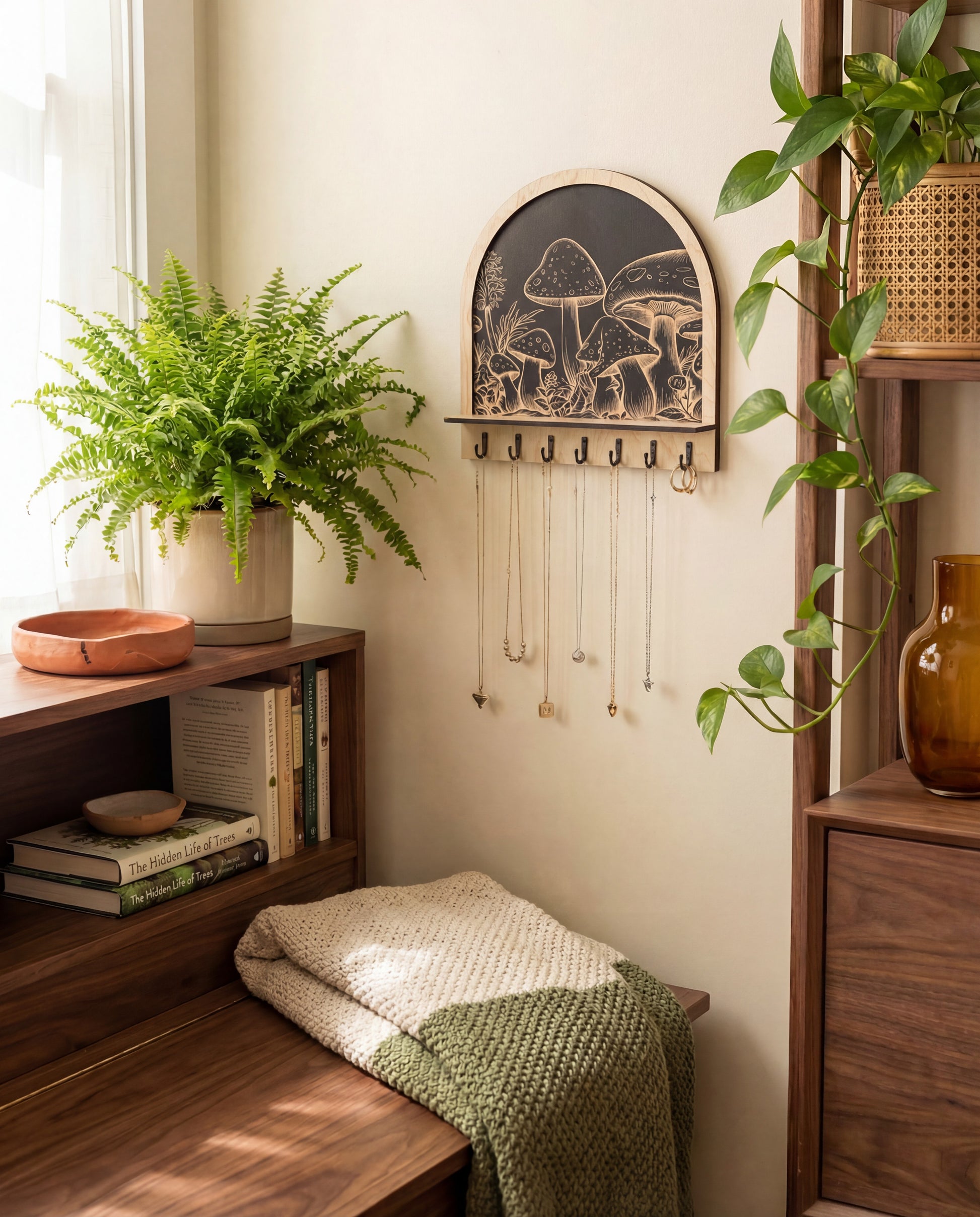 Arched wooden wall organizer with engraved mushroom artwork and black hooks, used as a key holder and jewelry hanger with necklaces, styled with plants and shelves.