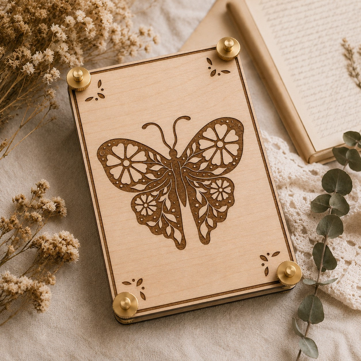 Handmade wooden flower press featuring a detailed laser-engraved butterfly with floral wing cutouts and decorative corner accents, displayed on a linen surface surrounded by dried wildflowers, eucalyptus sprigs, and an open book.