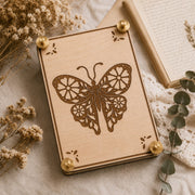 Handmade wooden flower press featuring a detailed laser-engraved butterfly with floral wing cutouts and decorative corner accents, displayed on a linen surface surrounded by dried wildflowers, eucalyptus sprigs, and an open book.