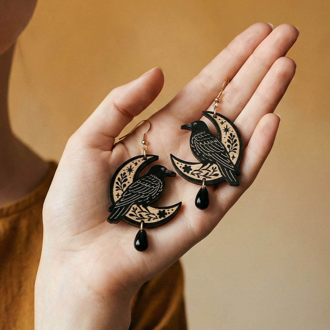 Hand holding decorative earrings with raven and moon design on a warm background.