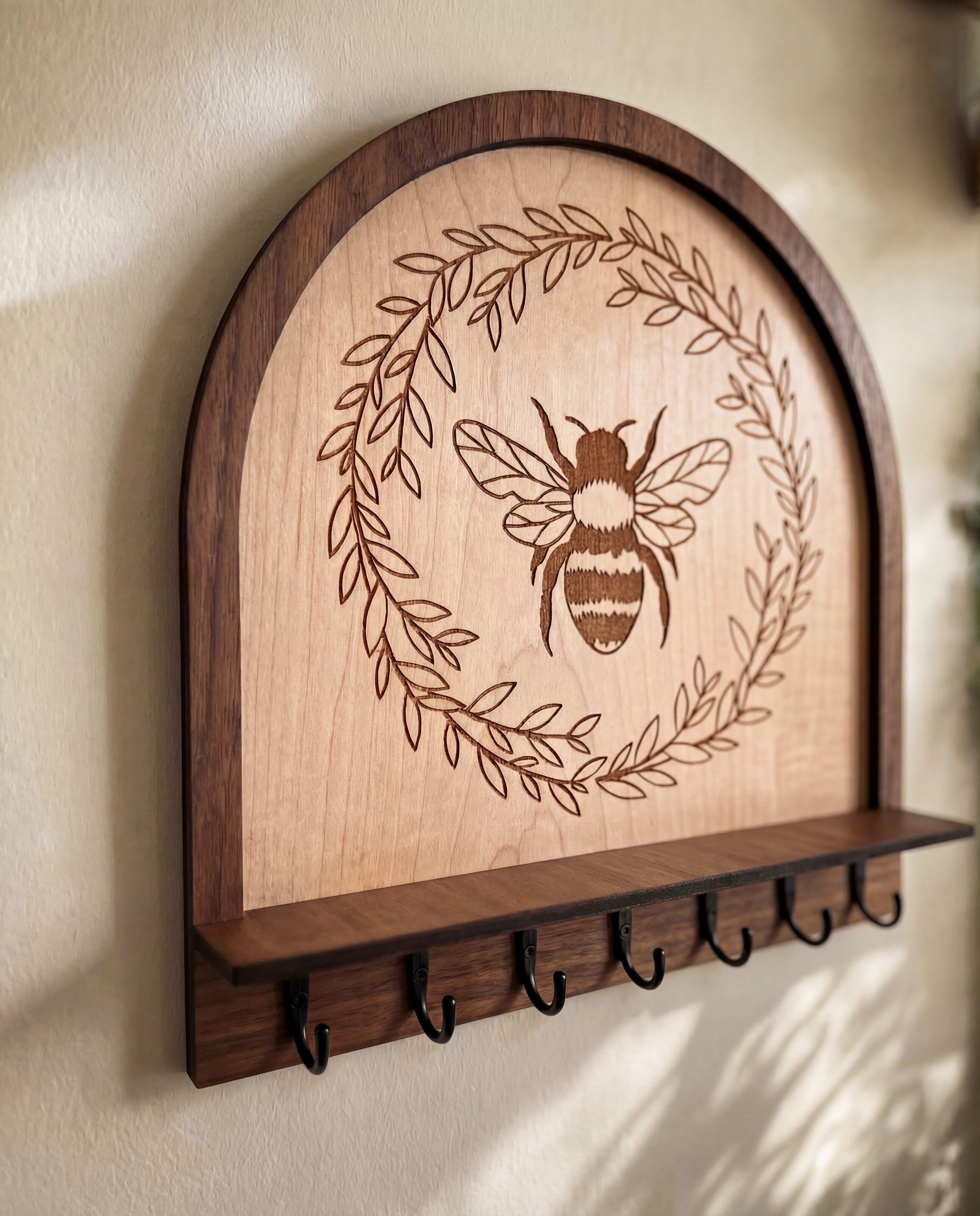 Close-up of maple wood bee wall organizer key holder with engraved honeybee and leafy wreath design, arched layered wood frame, small shelf ledge, and black metal hooks mounted on a neutral wall
