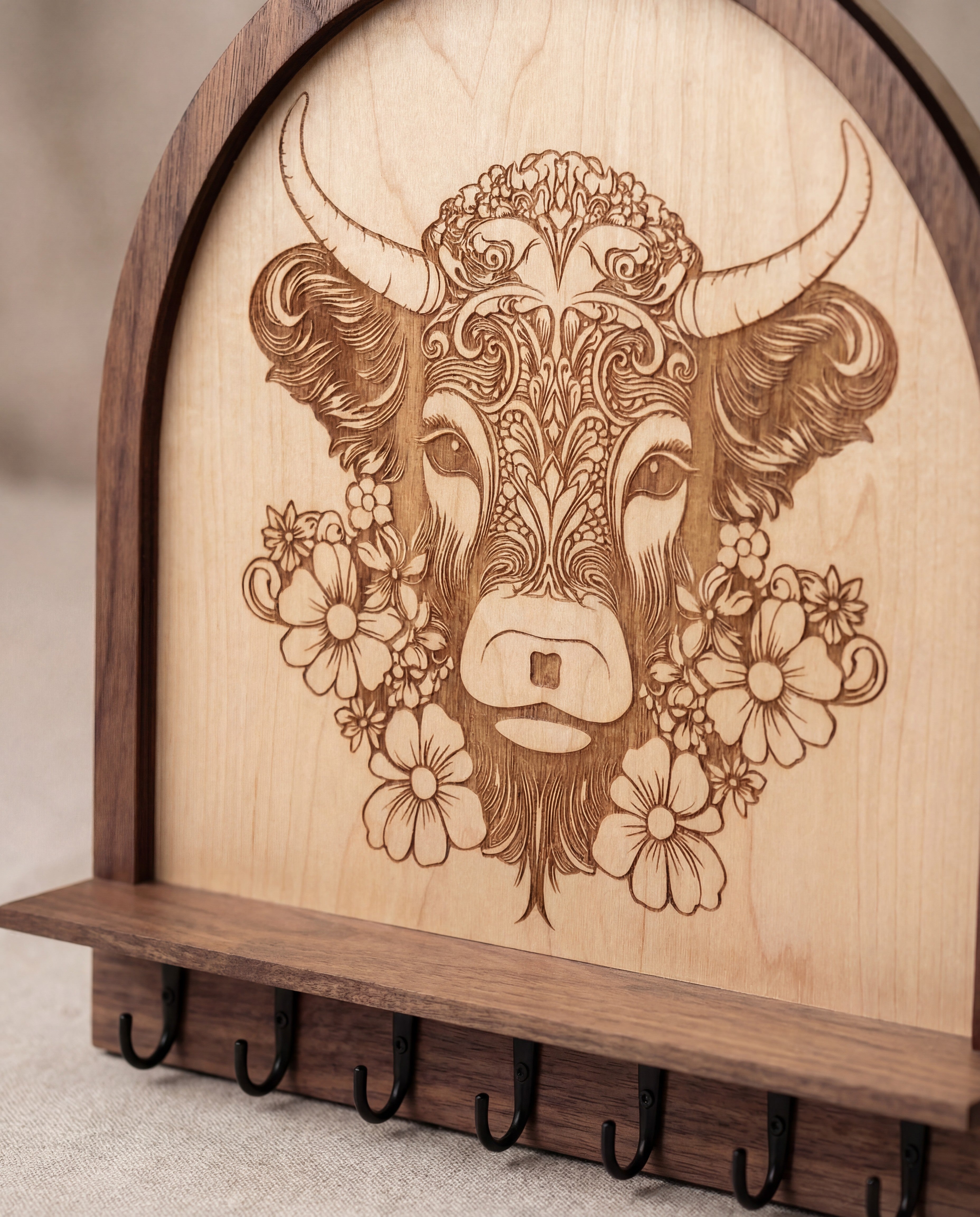 Macro close-up of the laser engraved floral highland cow artwork on a wooden wall key and jewelry holder, showing detailed engraving above a shelf with black metal hooks.