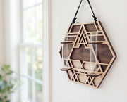 Geometric hexagon wall propagation station in layered maple wood with walnut-toned back layer and sacred geometry cutout design, hanging from a black suede strap and holding two clear glass vials, styled as modern boho wall decor by a bright window.
