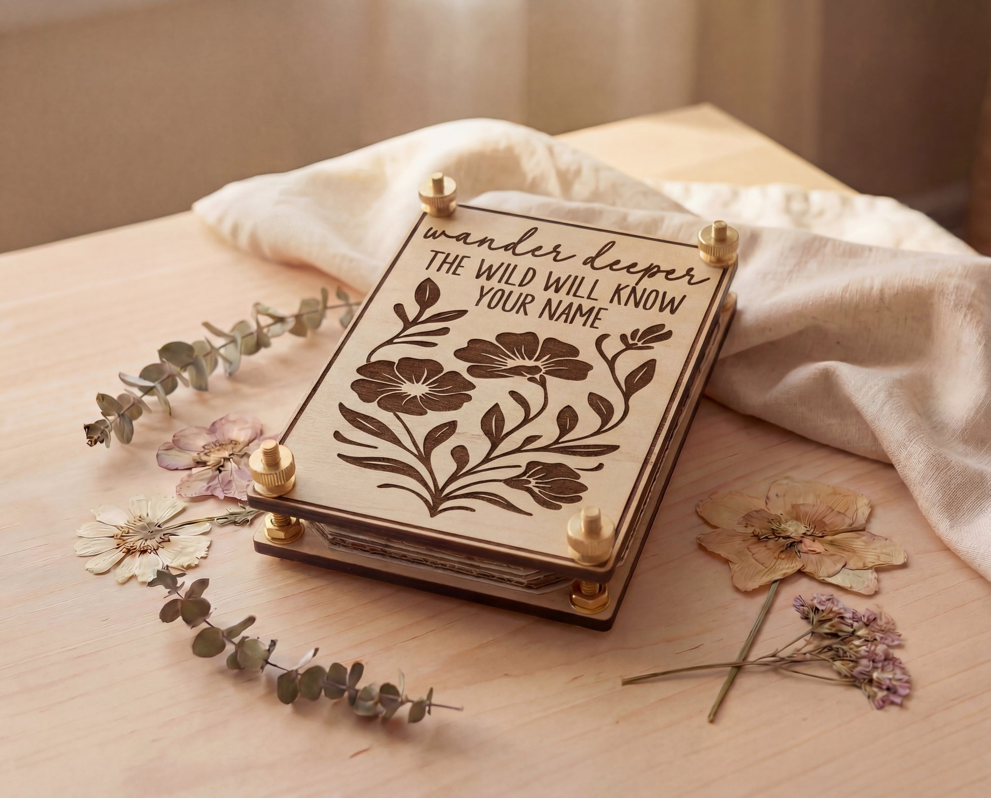 Handmade 4x6 maple flower press with brass hardware and “Wander deeper, the wild will know your name” engraved floral design, styled on a light wood table with linen cloth, eucalyptus sprigs, and pressed flowers.
