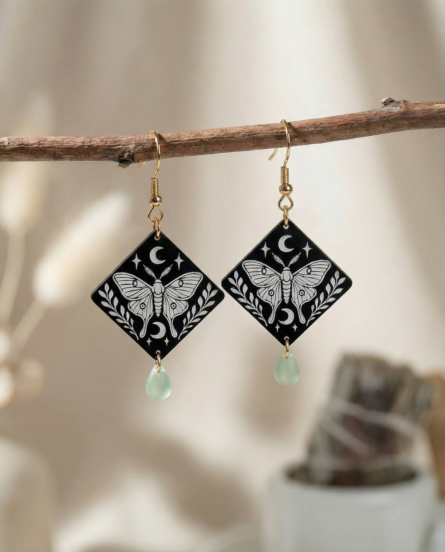 Engraved moth diamond-shaped wood dangle earrings with crescent moon details, mint Czech glass teardrop beads, and 14k gold hooks hanging on a branch.