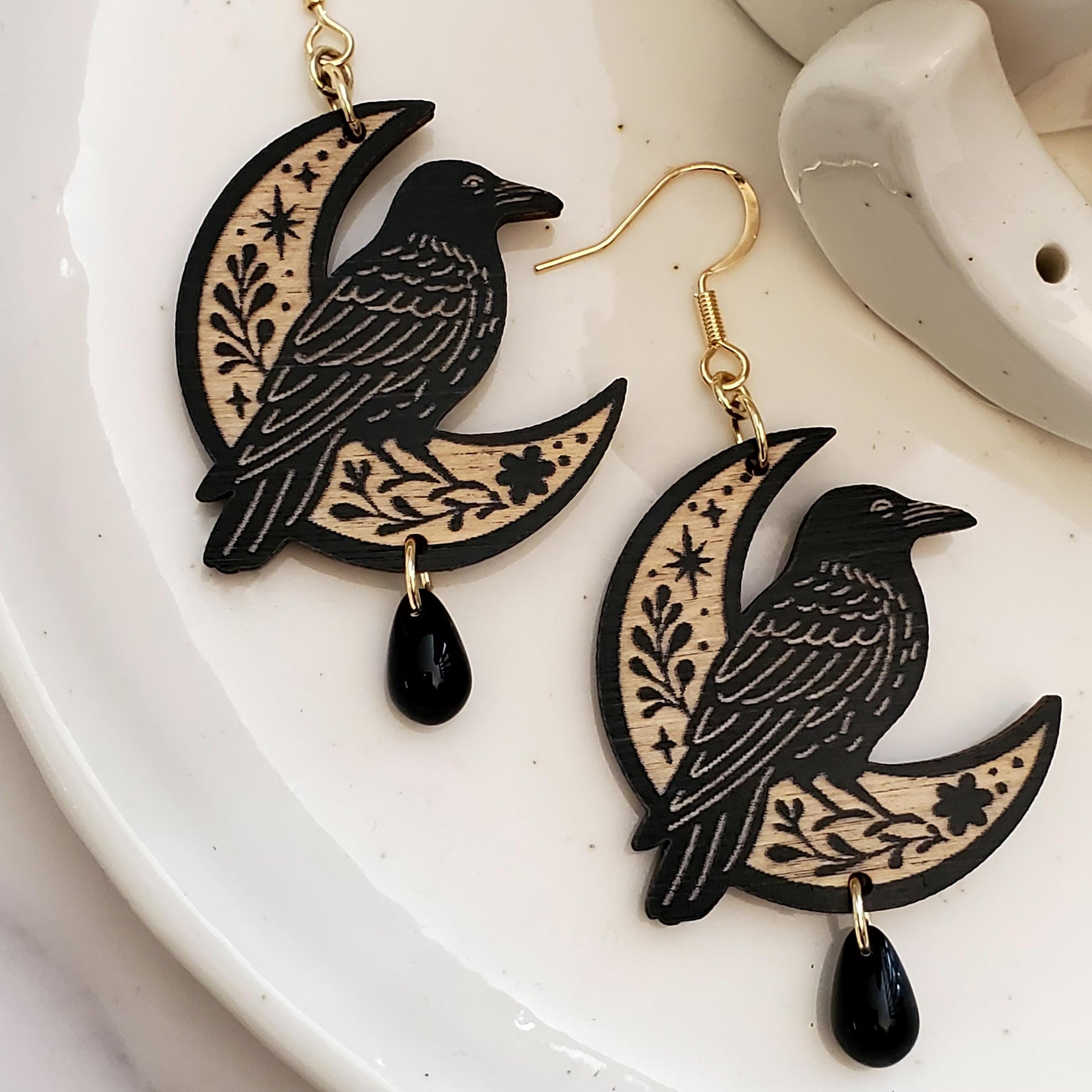 Black painted wooden earrings in the shape of a raven sitting on a moon with a floral engraving sitting in a white dish on a white surface.