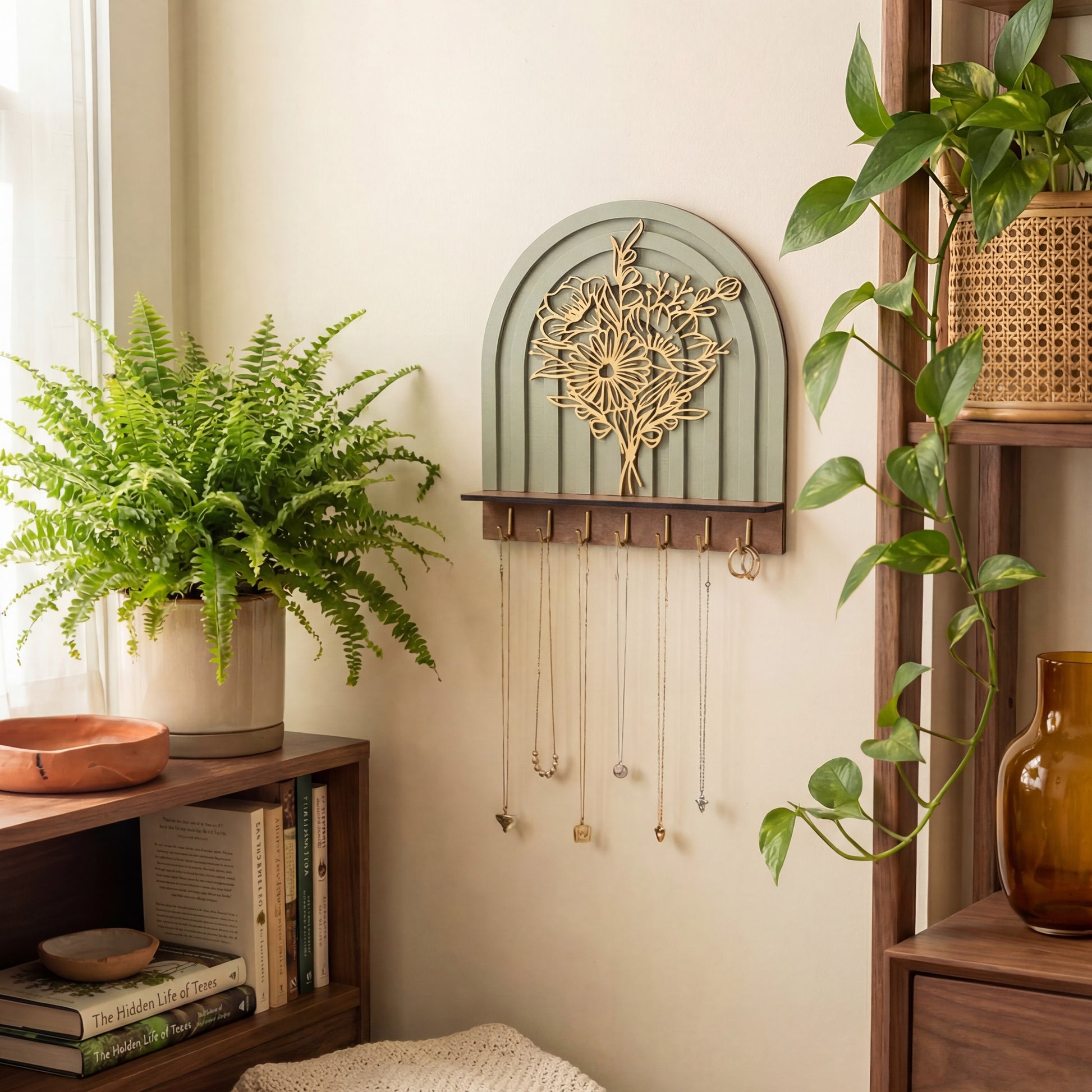 Arched wall-mounted jewelry organizer with layered floral botanical design, wood shelf, and brass hooks holding necklaces in a cozy boho room.