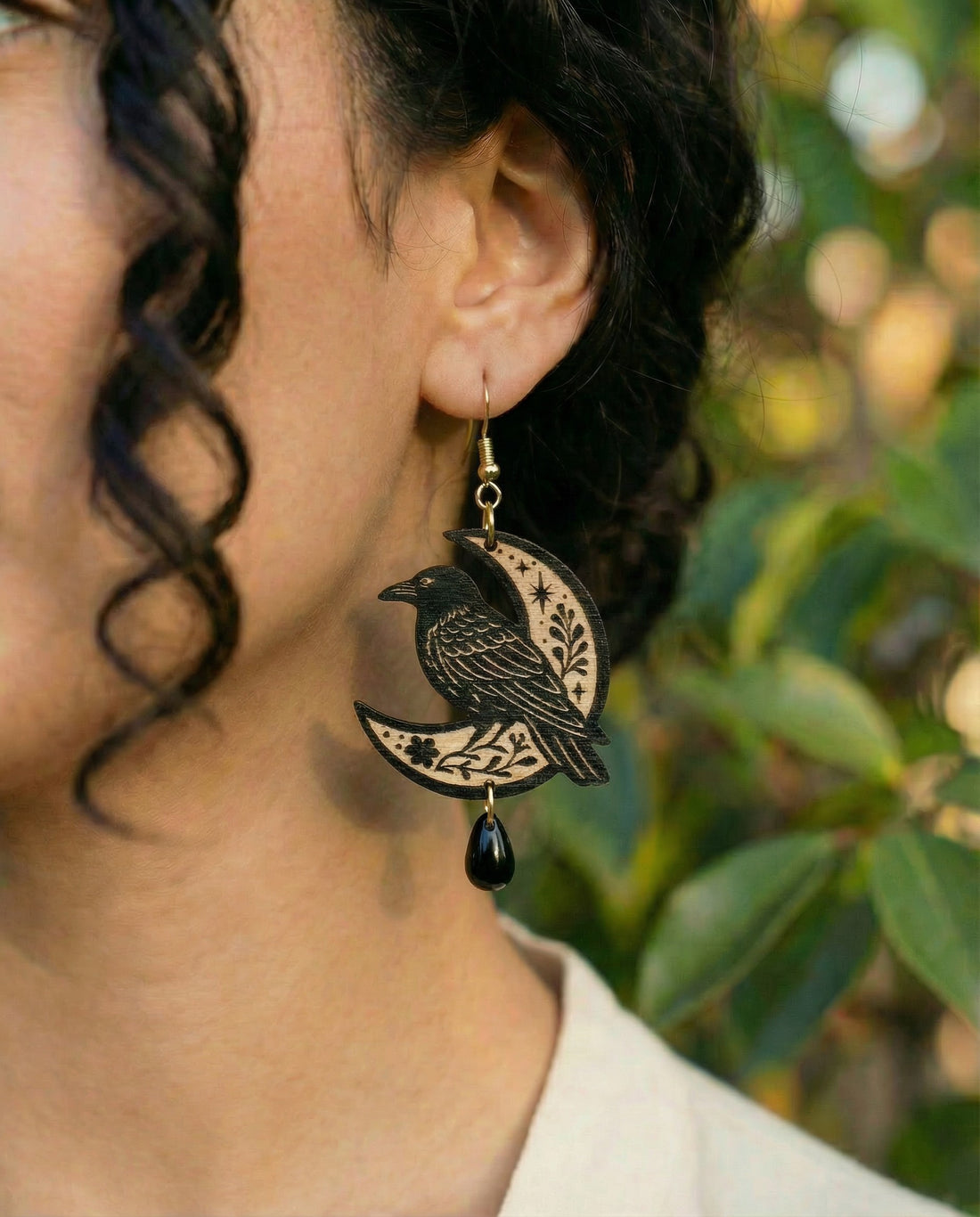 Earring with a raven sitting on a crescent moon worn by a person against a blurred natural background