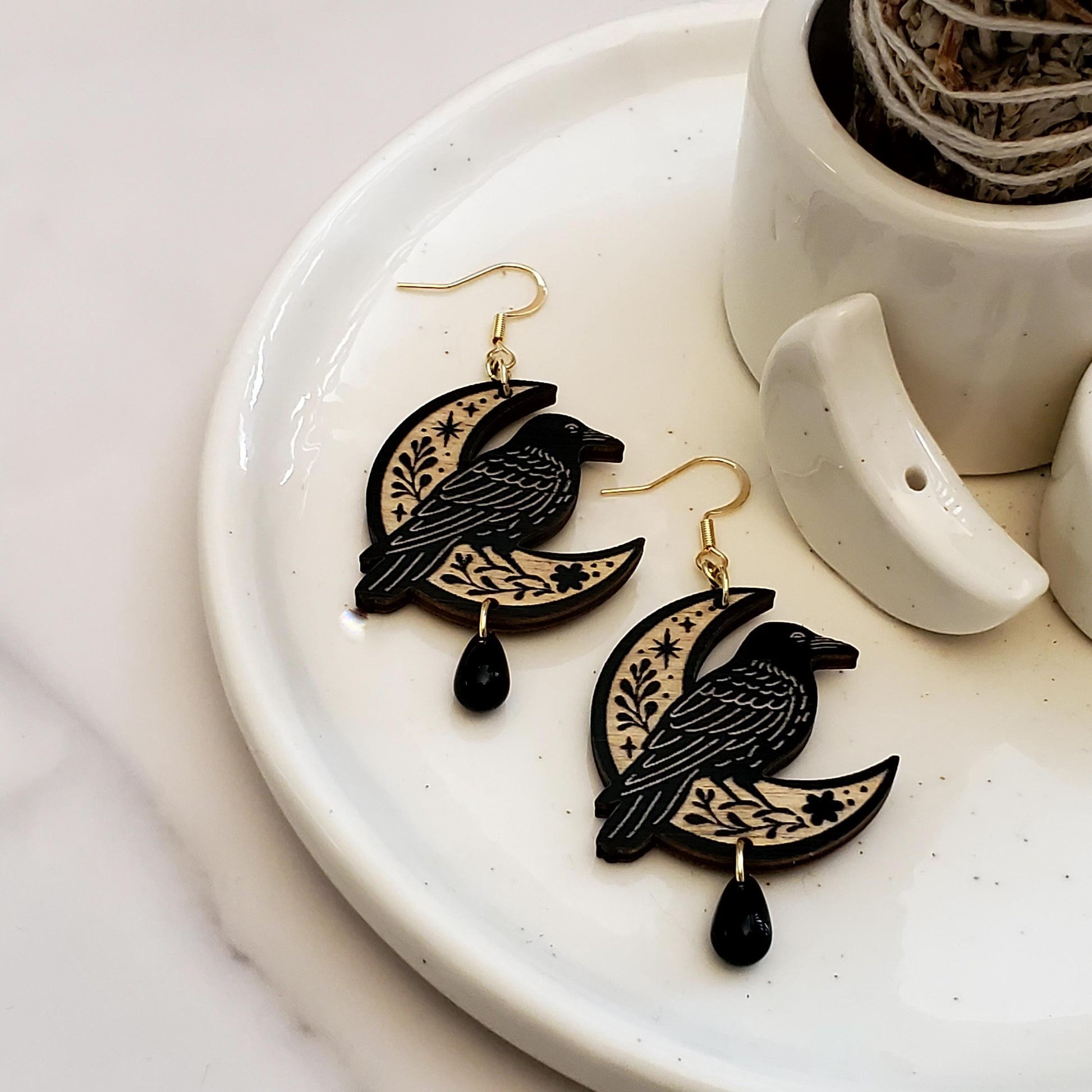 Black painted wooden earrings in the shape of a raven sitting on a moon with a floral engraving sitting in a white dish on a white surface.